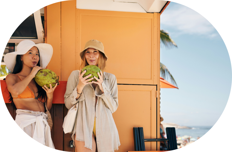 couple of woman drinking coconut water from coconuts with straws on the beach
