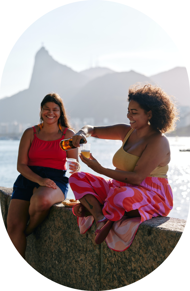 couple of woman drinking alcohol, one pours beer on a plastic cup, the other watches and smiles, they are sitting by the water, mountains in the background